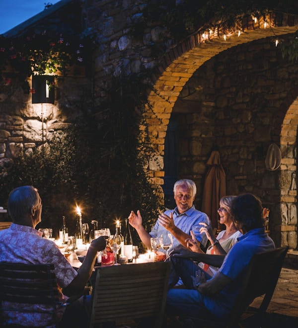 En gruppe voksne venner sitter rundt et utendørs spisebord og spiser og drikker. De snakker alle lykkelig og gleder seg over hverandres selskap når mørket faller på. Bildet er tatt i Toscana, Italia.