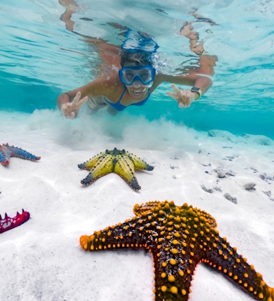 Voksen kvinne som viser fredsskilt mens hun snorkler rundt tropisk sjøstjerne.