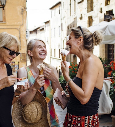 Tre venninner som står og spiser italiensk iskrem mens de står i en gate i Toscana om sommeren. De smiler og vender seg mot hverandre mens de nyter ferien.