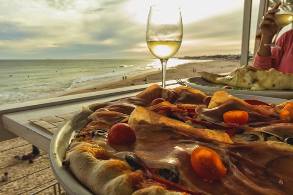 Hjemmelaget pizza med skinke, tomat, oliven og paprika med et glass hvitvin og stranden ved solnedgang i bakgrunnen