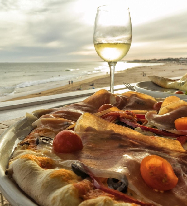 Hjemmelaget pizza med skinke, tomat, oliven og paprika med et glass hvitvin og stranden ved solnedgang i bakgrunnen