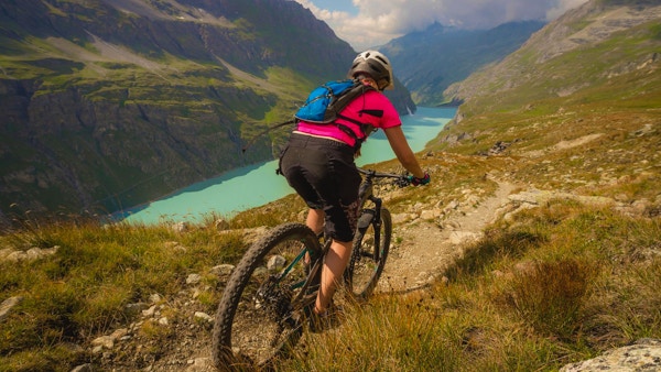 En dame med en rosa T-skjorte som sykler på terrengsykkel langs en løype over en blå innsjø i de sveitsiske alpene.