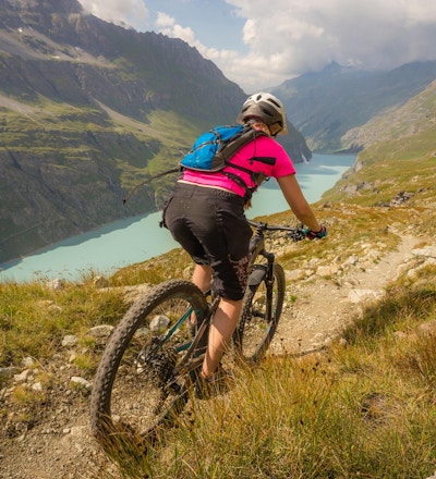 En dame med en rosa T-skjorte som sykler på terrengsykkel langs en løype over en blå innsjø i de sveitsiske alpene.