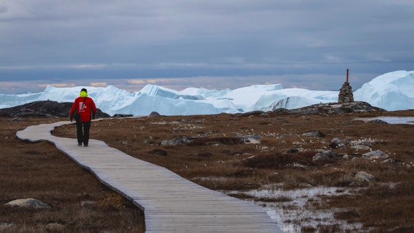 En rødkledd turist som vandrer på en sti, med store isfjell og grå himmel i bakgrunnen