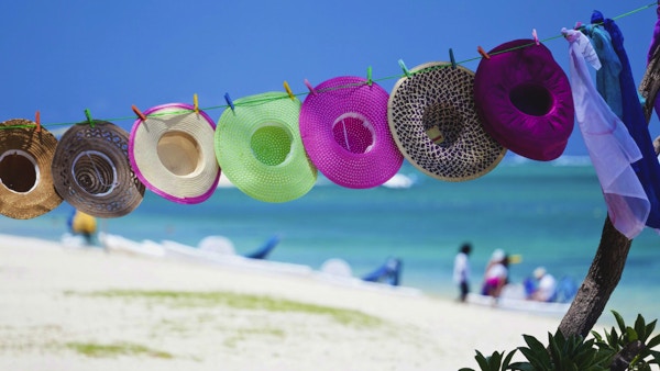 Detaljhandel på stranden, mauritansk stil. Halmhatter og strandomslag for salg på en tropisk strand på en varm, solrik dag i paradis, også kalt Mauritius. Turister i bakgrunnen som leier en pedalbåt for å padle i det turkise vannet i den naturlige lagunen.
