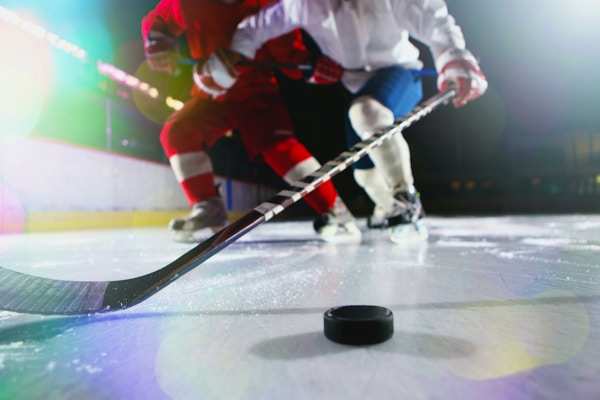 Isnivåbilde av mannlige ishockeyspillere i en takling under et spill. De høye hvite sokkene og de mørkeblå buksene til hockeyspilleren kan sees. Det er en hockeyspinne som strekker seg etter pucken. En hockeyspuck som kaster to skygger er på isen i høyre hjørne av bildet.