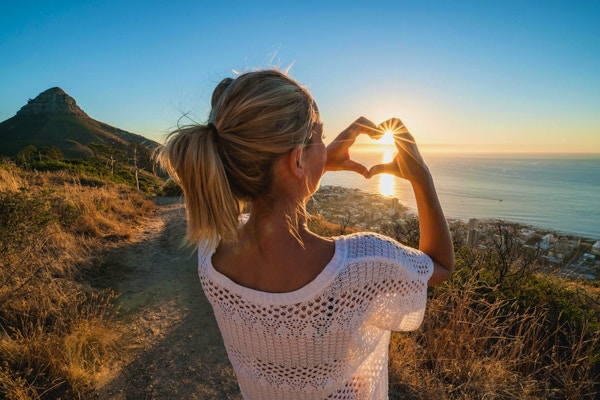 Ung kvinne ved sjøen som ser på solnedgang og lager en hjerteformet fingerramme. Skutt i Cape Town, Sør-Afrika.