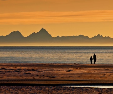 To mennesker i silhuett på stranda med fantastisk midnattssol med fjell i bakgrunnen.