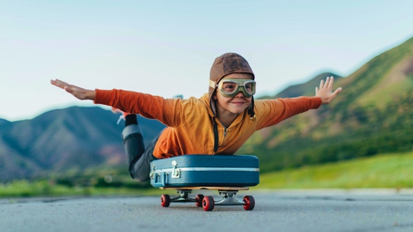 En ung gutt iført flybriller og flyhette har pakket kofferten og lagt den på et skateboard. Han er klar til å fly bort med armene utstrakt til drømmemålet. Bildet er tatt i Utah, USA.