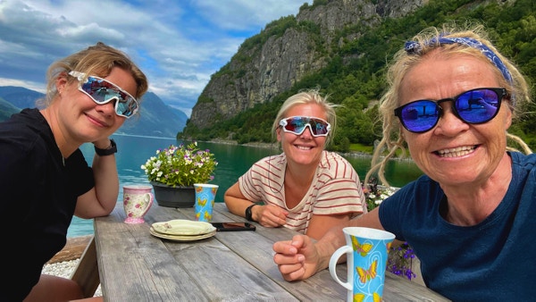 Tre damer ved trebord og kaffekopper med hav og fjell i bakgrunnen.
