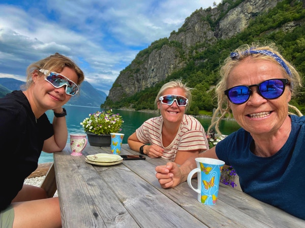 Tre damer ved trebord og kaffekopper med hav og fjell i bakgrunnen.