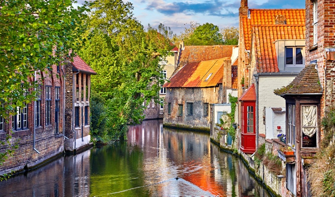 Brugge, Belgia. Middelalderske gamle hus laget av gamle murstein ved vannkanal med båter i gamlebyen. Sommersolnedgang med solskinn og grønne trær. Pittoreske landskap.