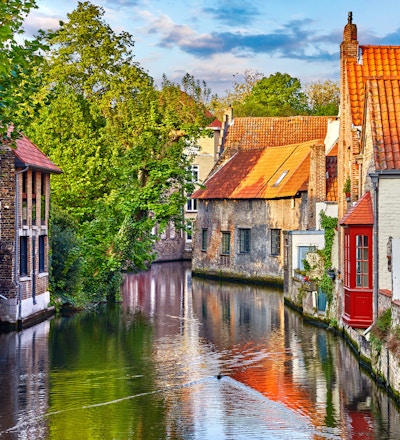 Brugge, Belgia. Middelalderske gamle hus laget av gamle murstein ved vannkanal med båter i gamlebyen. Sommersolnedgang med solskinn og grønne trær. Pittoreske landskap.