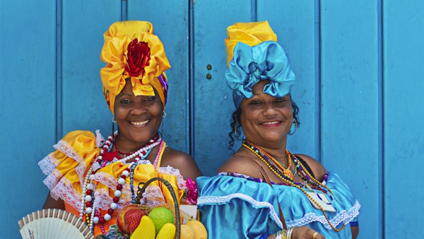 Portrett av glade cubanske kvinner som står mot blå trevegg. Smilende modne kvinner er i tradisjonelle kjoler. De er med fruktkurv og håndvifte.