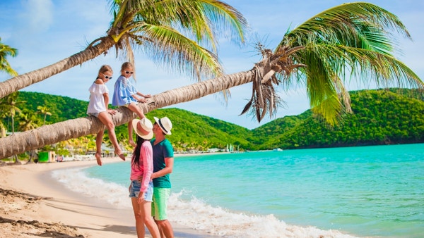 To jenter sitter på en palme med foreldrene ved siden av på sandstrand.