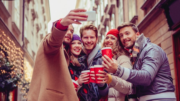 Venner tar selfies utendørs i vinterbygate. Iført varme klær, luer og skjerf. Holder kaffe å gå. Wien, Østerrike. Festlig lysslynge i bakgrunnen.