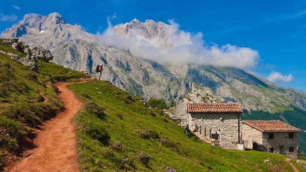 En turgåermann som stiger opp på klatreruten til det emblematiske Picu Urriellu (Naranjo de Bulnes), og beundrer den imponerende utsikten over grønne daler og steinete fjell. Det er en liten gruppe beitehus og storfehytter, og også et lite fjelltilfluktssted for gjetere og fjellklatrere. Refuge of The Tenerosa, Picos de Europa, Asturias, Spania