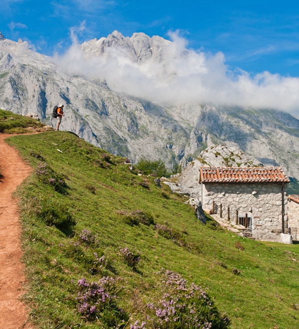 En turgåermann som stiger opp på klatreruten til det emblematiske Picu Urriellu (Naranjo de Bulnes), og beundrer den imponerende utsikten over grønne daler og steinete fjell. Det er en liten gruppe beitehus og storfehytter, og også et lite fjelltilfluktssted for gjetere og fjellklatrere. Refuge of The Tenerosa, Picos de Europa, Asturias, Spania