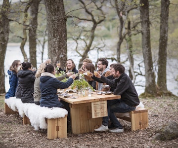 Gruppe menneske sitter ved utendørs langbord om vpren og spiser og drikker.
