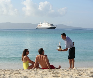 Mannskap tilbyr drikke og frukt til par på stranden og yachten ligger på havet i bakgrunnen