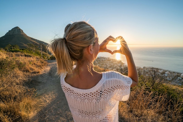 Ung kvinne ved sjøen som ser på solnedgang og lager en hjerteformet fingerramme. Skutt i Cape Town, Sør-Afrika.