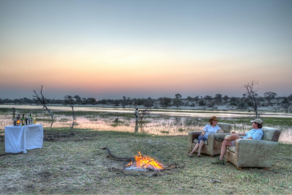 Par i lenestoler med drinker i afrikansk landskap med leirbål i solnedgang.