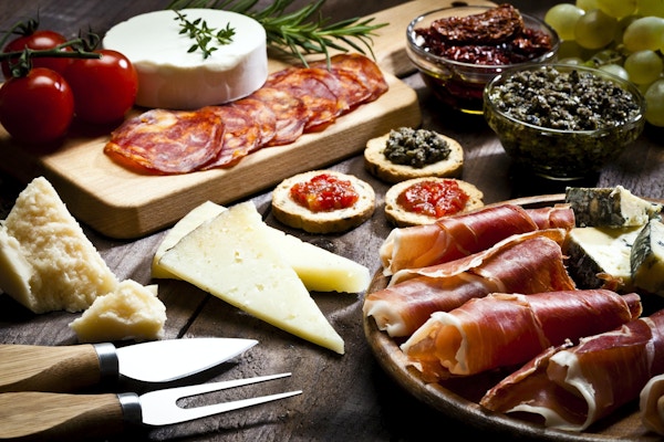 Rustikk trebord fylt med et utvalg av god mat. Et trebrett med prosciutto og ost er nederst til høyre på rammen. To glassboller med tørkede tomater og olivenpasta og et skjærebrett er øverst.