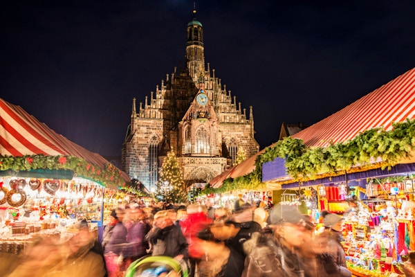 En stor folkemengde går gjennom Nürnbergs verdensberømte julemarked (Christkindlsmarkt) om kvelden, mens de passerer fargerike juledekorasjoner og matboder. Nürnbergs landemerke Frauenkirche (Church of our Lady) sees i bakgrunnen.