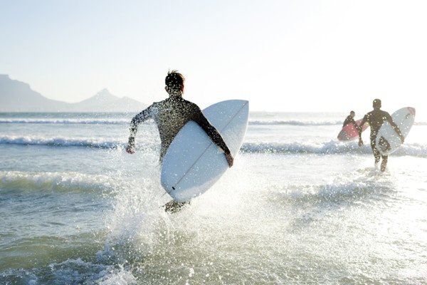 Tre tenåringsgutter som har på seg våtdraktene sine med surfebrettene, løper ut i havet på en solrik sommerdag.