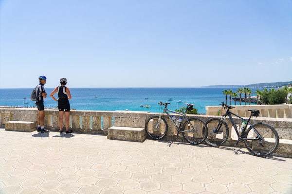 To syklister med hjelm står ved siden av sine parkerte sykler og ser utover havet