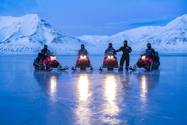 Fire snøscootere med sjåfører på isen med fjell i polarlys i bakgrunnen