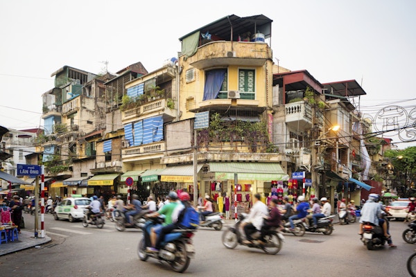 Opptatt gatehjørne i gamlebyen i Hanoi, Vietnam. Mange mennesker pendler på motorsykler eller biler. Gaten er omgitt av butikker og leilighetsbygg.