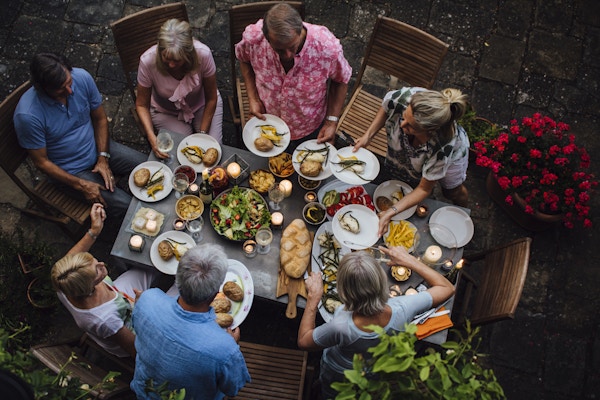 En gruppe voksne venner sitter rundt et utendørs spisebord og spiser og drikker. De snakker alle lykkelig og gleder seg over hverandres selskap. Bildet er tatt ovenfra, i Toscana, Italia.