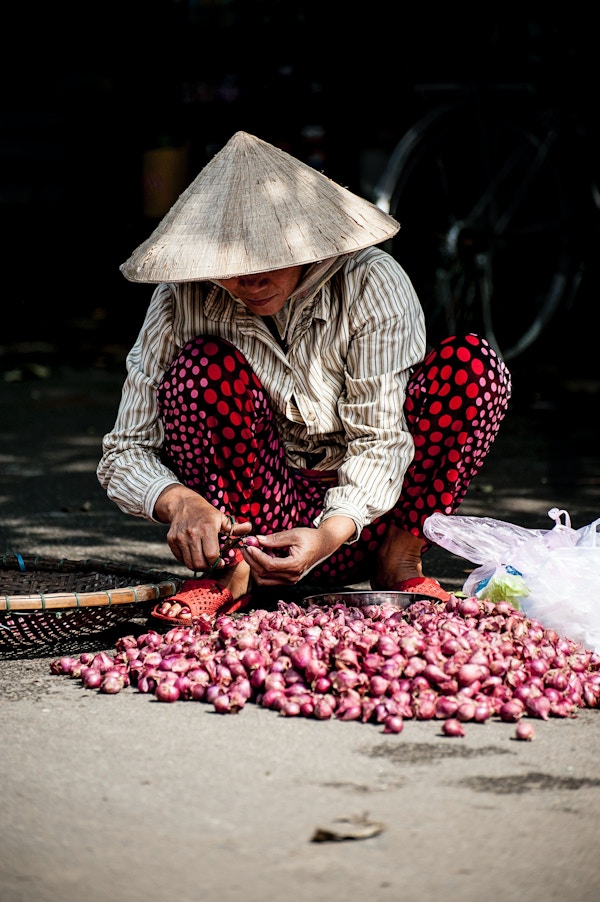 Kvinne sitter på huk og kutter til små lilla løk på et matmarked