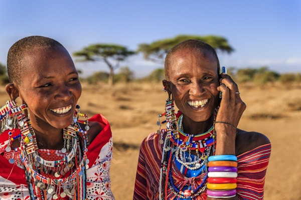 To masai-kvinner i fargerike klær og pynt, den ene snakker i mobiltelefon.