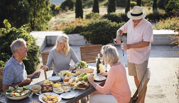 En gruppe voksne venner sitter rundt et utendørs spisebord, spiser og drikker. De feirer ferien med et glass champagne og koser seg med hverandres selskap. Bildet er tatt i Toscana, Italia.