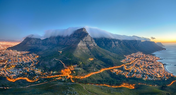 Table Mountain dekket av sitt typiske skylag og De Tolv Apostles. Nedenfor på venstre side kan man se Cape Town og den vakre Campsbukten på høyre side. Bildet er tatt rett etter solnedgang.