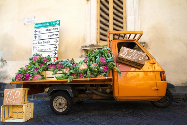 En søt oransje vintagebil som selger lilla blomkål på en gate i Sicilia, Italia. Gul vegg, skodder og veiskilt i bakgrunnen.