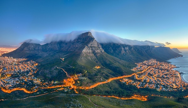 Table Mountain dekket av sitt typiske skylag og De Tolv Apostles. Nedenfor på venstre side kan man se Cape Town og den vakre Campsbukten på høyre side. Bildet er tatt rett etter solnedgang.