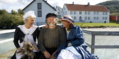 Mann og to kvinner kledd i gammeldagse kjær sittende på brygga med historiske bygninger i bakgrunnen.