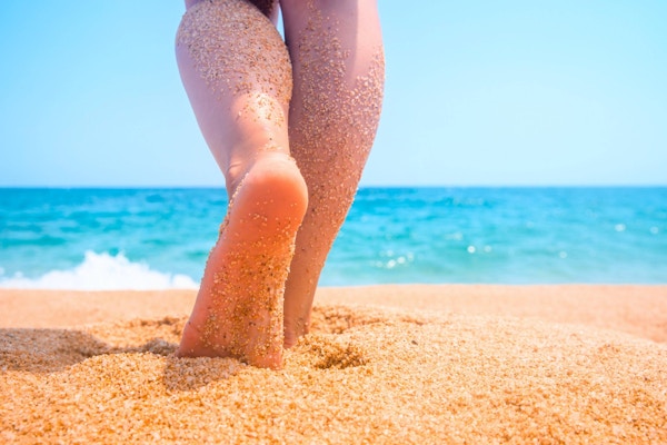 Tropisk strandbakgrunn. Feriekonsept. Slanke kvinnelige ben med sand på en blå sjøbakgrunn. Tropisk feriested.