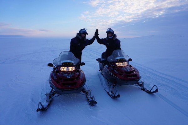To personer kjører hver sin snøskuter i solnedgang