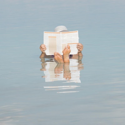 Kvinne med kryssede føtter, holder og leser en avis mens hun flyter i salt vann i Dødehavet, Israel, Midtøsten.