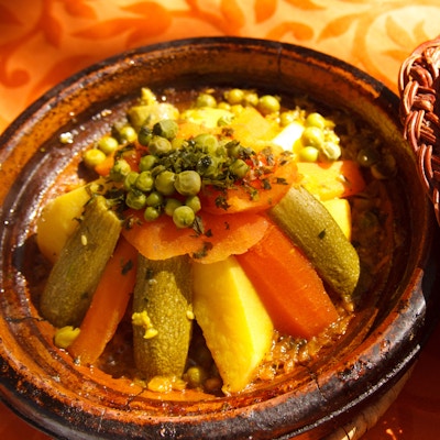 En pen vegetabilsk tagine på en sprukket leirplate (den nederste halvdelen av tajine-gryten) ved siden av en kurv med brød. Bildet er tatt på en tradisjonell marokkansk restaurant i Zagora.