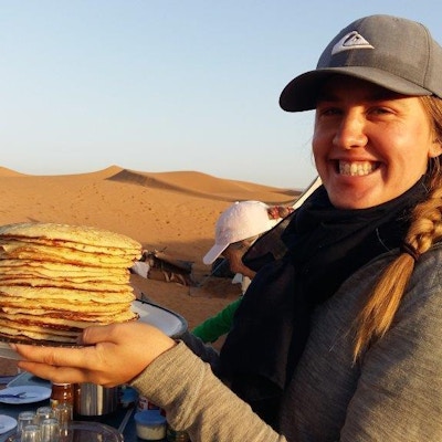 En dame holder et fat pannekaker over frokostbordet i ørkenen. Turdeltagere spiser frokost.