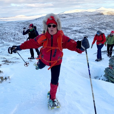 Kvinne med anorakk, truger og staver poserer på toppen av fjell.