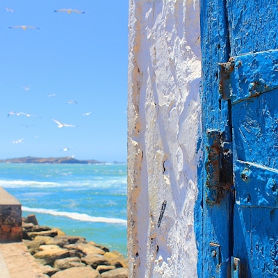 Deler av en blå dør og hvit murvegg i forgrunnen, hav i bakgrunnen.