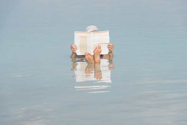 Kvinne med kryssede føtter, holder og leser en avis mens hun flyter i salt vann i Dødehavet, Israel, Midtøsten.