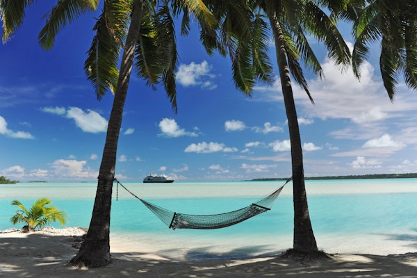 Et cruiseskip dukker opp i horisonten til en tropisk turkis lagune - med en hengekøye i halvsilhuett, skyggelagt av palmer