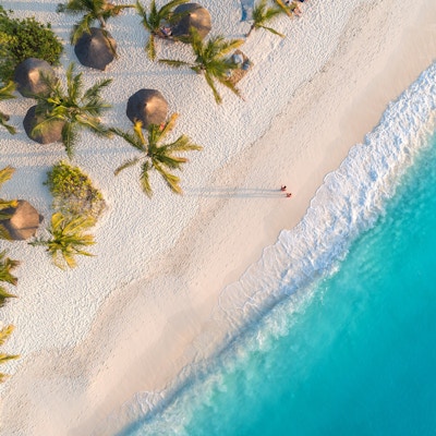 Luftfoto av parasoller, palmer på sandstranden i Indiahavet ved solnedgang. Sommerferie i Zanzibar, Afrika. Tropisk landskap med palmer, parasoller, hvit sand, blått vann, bølger. Topp utsikt